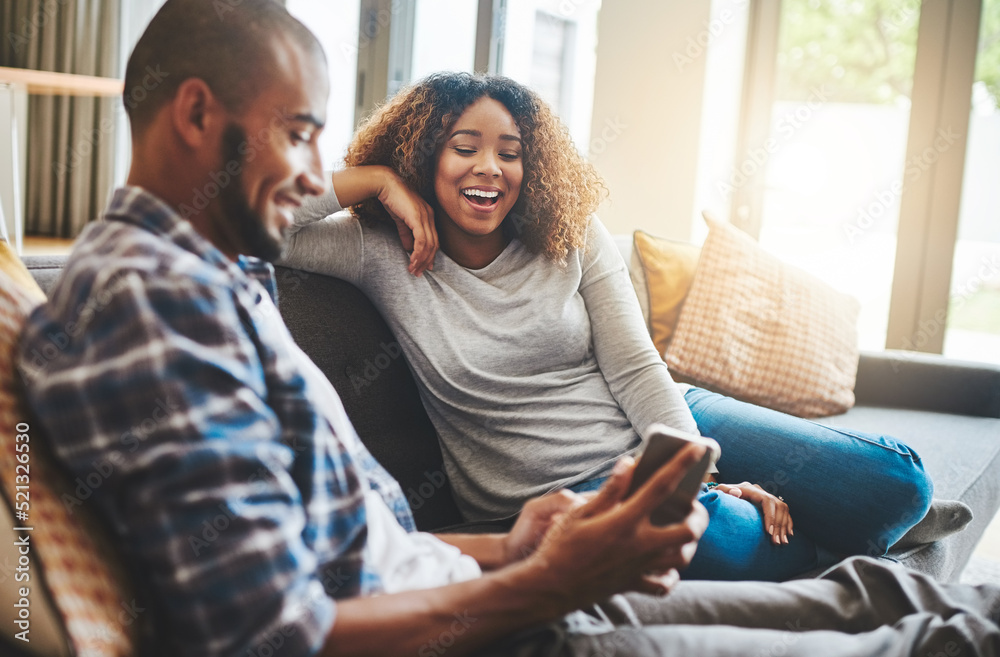 Obraz premium Happy, laughing and cheerful couple relaxing together and looking at social media on a phone at home while sitting on the sofa inside. Young husband and wife enjoying browsing online on a sunny day
