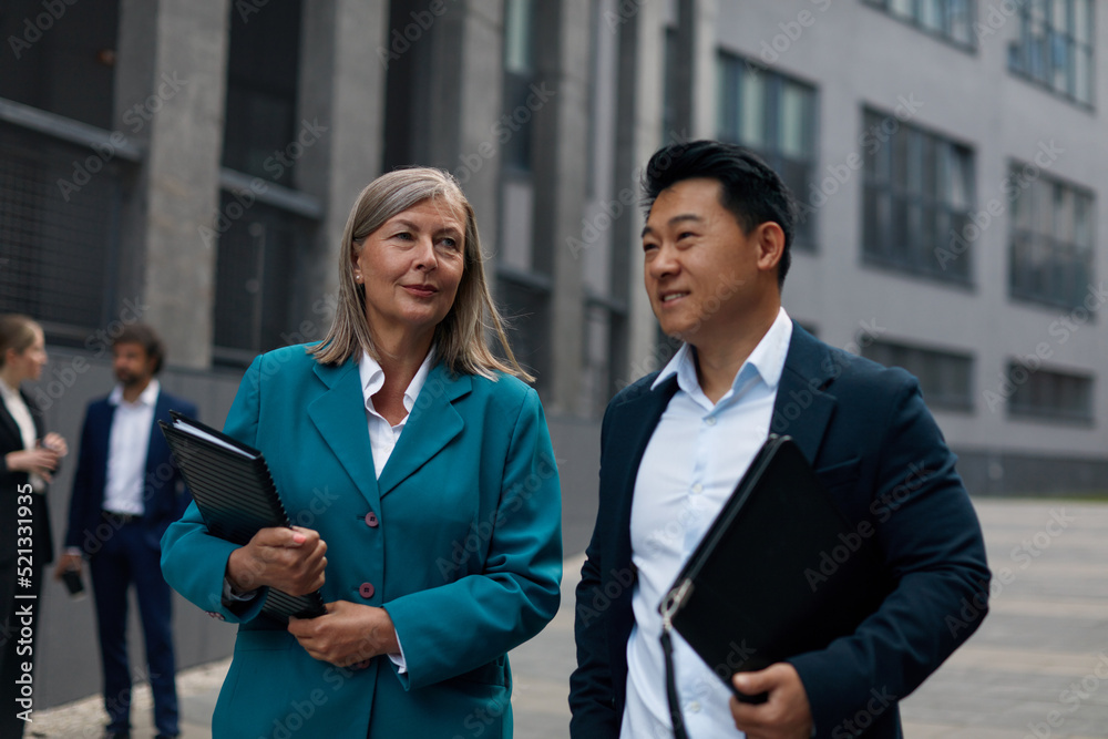 Man and Woman Colleagues Walking Discussing Documents. Colleagues in Elegant Suits Going on Street with Clipboard. Urban Background Man and Woman Colleagues Walking Discussing Documents. Colleagues in Elegant Suits Going on Street with Clipboard. Urban Background