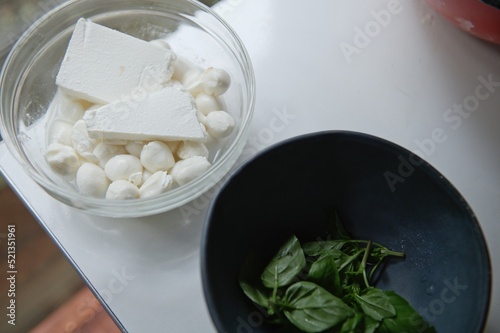 Top view of fresh ingredients: mozzarella and salted feta cheese in a glass transparent bowl, aromatic basil leaves on navy ceramic bowl on white kitchen table. Copy ad space. Food, cuisine, culinary