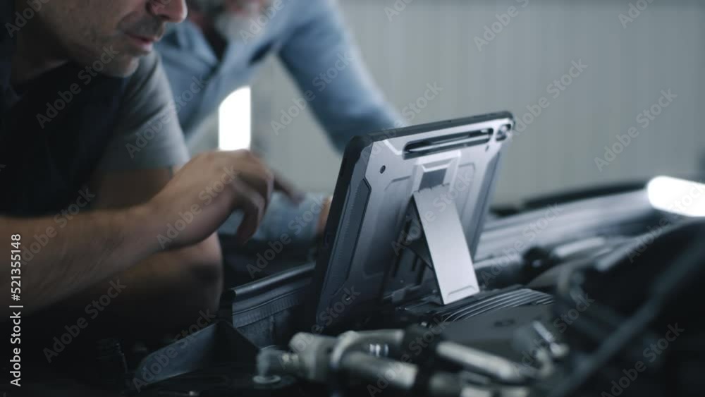 Car service mechanic in repair shop clicking on a screen of digital tablet computer while standing next to a client, near the opened bonnet of the car during diagnostics and maintenance Car service mechanic in repair shop clicking on a screen of digital tablet computer while standing next to a client, near the opened bonnet of the car during diagnostics and maintenance