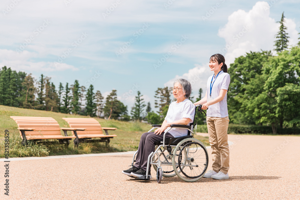 公園を散歩する介護士と車椅子の高齢者女性（青空）
