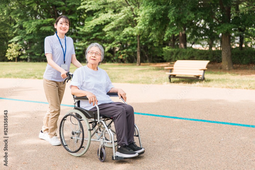 公園を散歩する介護士と車椅子の高齢者女性（緑）
