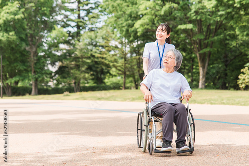 公園を散歩する介護士と車椅子の高齢者女性（緑） 