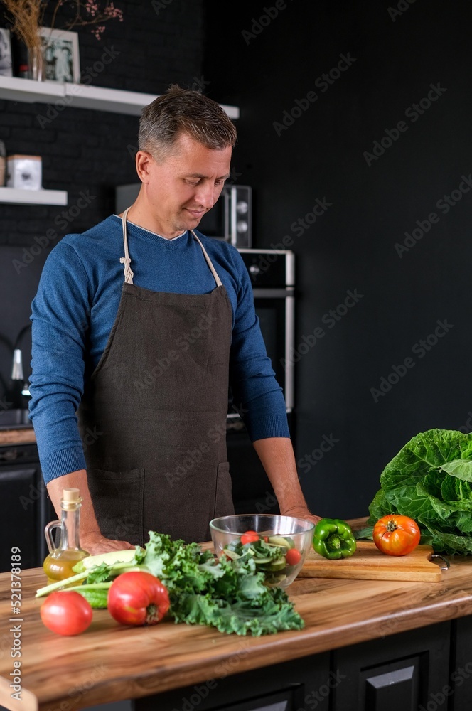 Smiling man cooking salad, cutting fresh vegetables in modern black kitchen, happy satisfied male preparing vegetarian food for dinner. Cabbage, tomatoes, zucchini, pepper on the back. Smiling man cooking salad, cutting fresh vegetables in modern black kitchen, happy satisfied male preparing vegetarian food for dinner. Cabbage, tomatoes, zucchini, pepper on the back.