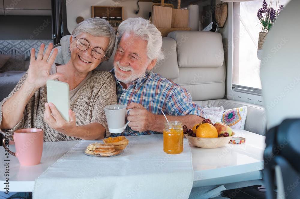 Happy senior couple sitting inside a camper van in video call with mobile phone, elderly retirees in alternative lifestyle enjoying breakfast together