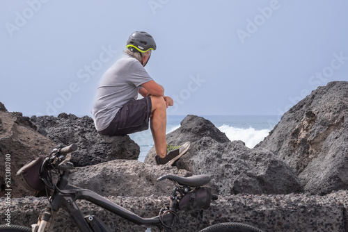 Senior man with helmet sitting on the rocks along the sea after running with his electric bicycle. Caucasian cyclist relaxing looking at the ocean waves photo