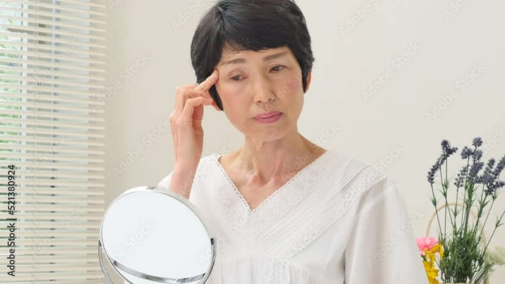 リビングで鏡で目元を見て悩むシニア女性 リビングで鏡で目元を見て悩むシニア女性