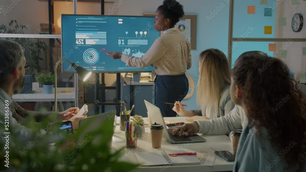Office worker pointing at digital screen and discussing marketing ideas in boardroom at night. Young lady making presentation for creative team.