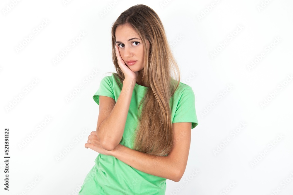 Very bored young beautiful woman wearing green T-shirt over white background holding hand on cheek while support it with another crossed hand, looking tired and sick, Very bored young beautiful woman wearing green T-shirt over white background holding hand on cheek while support it with another crossed hand, looking tired and sick,