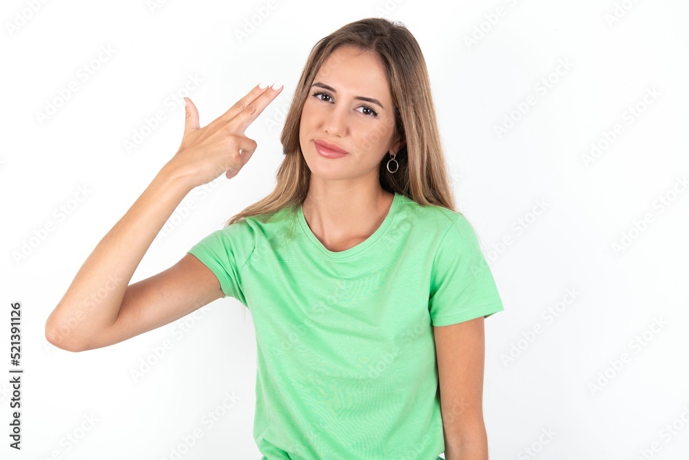 Unhappy young beautiful woman wearing green T-shirt over white background makes suicide gesture and imitates gun with hand, curves lips, keeps two fingers on temple Unhappy young beautiful woman wearing green T-shirt over white background makes suicide gesture and imitates gun with hand, curves lips, keeps two fingers on temple