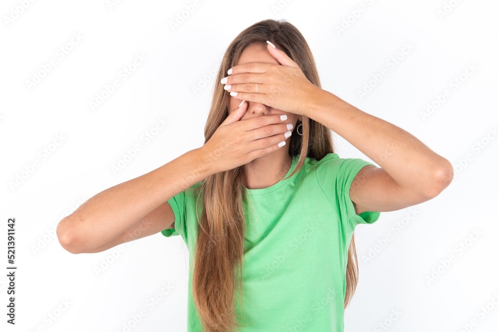 young beautiful woman wearing green T-shirt over white background Covering eyes and mouth with hands, surprised and shocked. Hiding emotions. young beautiful woman wearing green T-shirt over white background Covering eyes and mouth with hands, surprised and shocked. Hiding emotions.