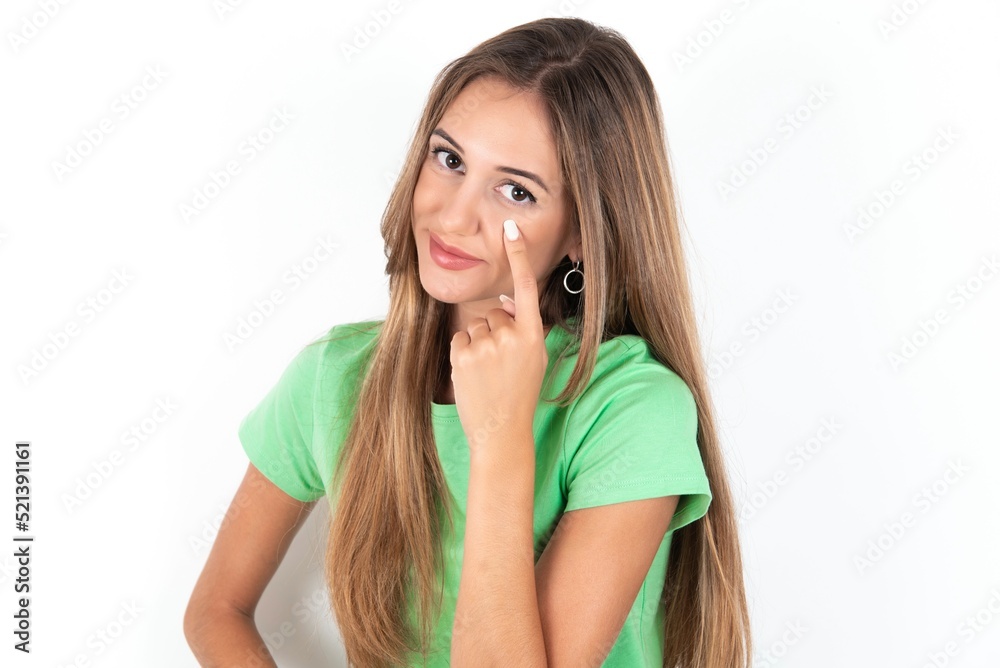 young beautiful woman wearing green T-shirt over white background Pointing to the eye watching you gesture, suspicious expression. young beautiful woman wearing green T-shirt over white background Pointing to the eye watching you gesture, suspicious expression.