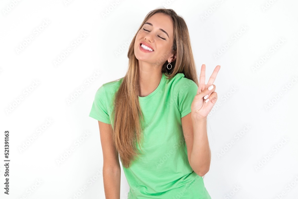 young beautiful woman wearing green T-shirt over white background smiling with happy face winking at the camera doing victory sign. Number two. young beautiful woman wearing green T-shirt over white background smiling with happy face winking at the camera doing victory sign. Number two.
