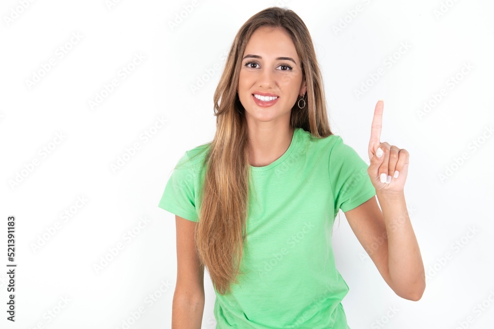 young beautiful woman wearing green T-shirt over white background showing and pointing up with finger number one while smiling confident and happy. young beautiful woman wearing green T-shirt over white background showing and pointing up with finger number one while smiling confident and happy.