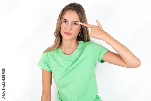 Unhappy young beautiful woman wearing green T-shirt over white background makes suicide gesture and imitates gun with hand, curves lips keeps two fingers on temple, shoots, being tired of everything,