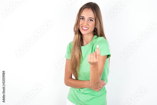 young beautiful woman wearing green T-shirt over white background Beckoning come here gesture with hand inviting welcoming happy and smiling