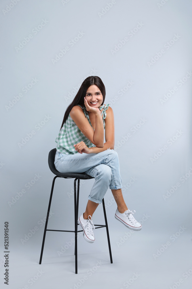 Beautiful young woman sitting on stool against light grey background Beautiful young woman sitting on stool against light grey background