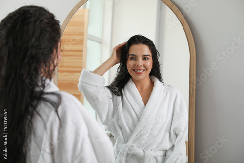 Beautiful young woman in bathrobe standing near mirror indoors