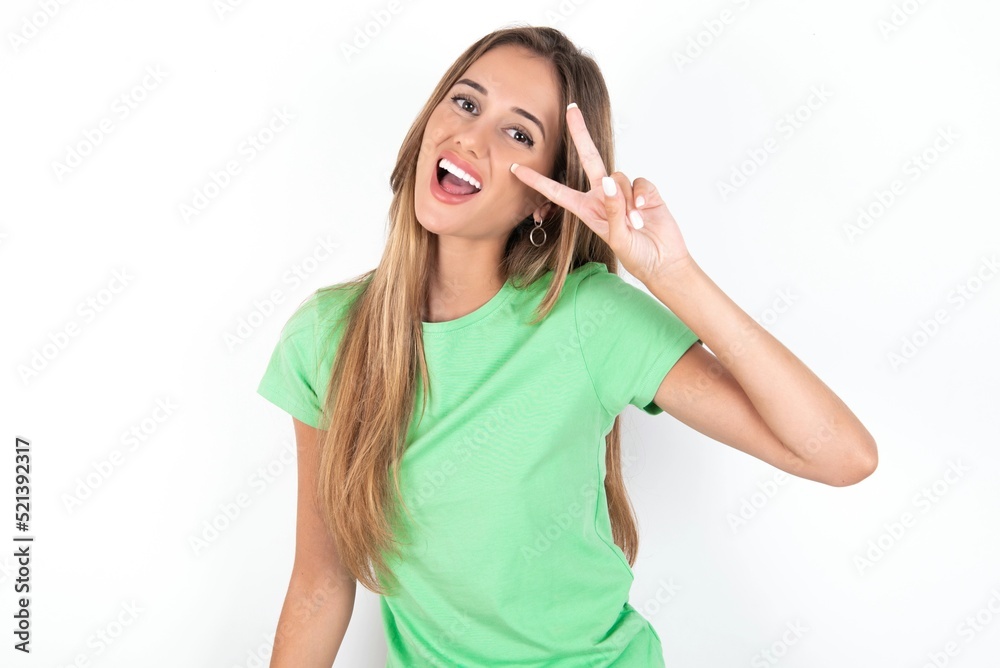 young beautiful woman wearing green T-shirt over white background, Doing peace symbol with fingers over face, smiling cheerful showing victory young beautiful woman wearing green T-shirt over white background, Doing peace symbol with fingers over face, smiling cheerful showing victory