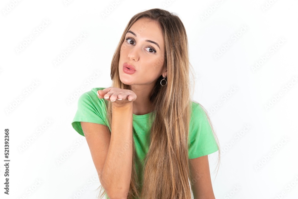 young beautiful woman wearing green T-shirt over white background, looking at the camera blowing a kiss with hand on air being lovely and sexy. Love expression. young beautiful woman wearing green T-shirt over white background, looking at the camera blowing a kiss with hand on air being lovely and sexy. Love expression.