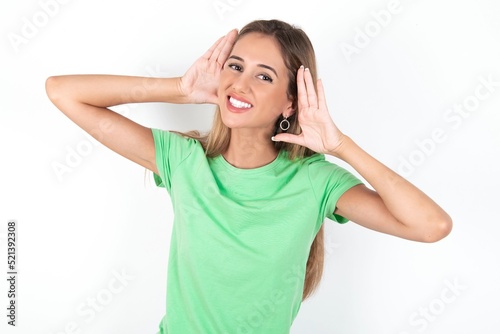 young beautiful woman wearing green T-shirt over white background, Trying to hear both hands on ear gesture, curious for gossip. Hearing problem, deaf © Roquillo