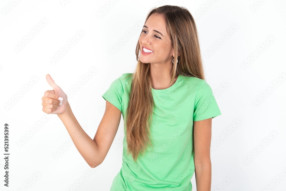 young beautiful woman wearing green T-shirt over white background, Looking proud, smiling doing thumbs up gesture to the side. Good job! young beautiful woman wearing green T-shirt over white background, Looking proud, smiling doing thumbs up gesture to the side. Good job!