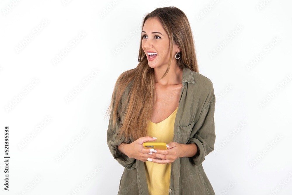 young beautiful woman wearing green overshirt over white background holding a smartphone and looking sideways at blank copyspace. young beautiful woman wearing green overshirt over white background holding a smartphone and looking sideways at blank copyspace.