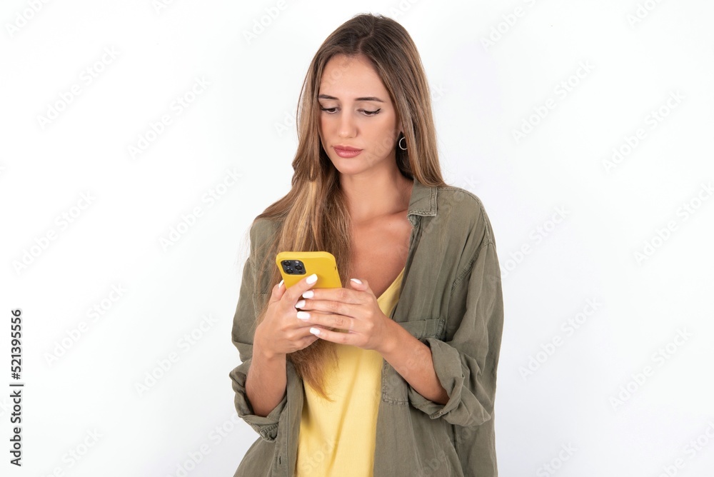 Excited young beautiful woman wearing green overshirt over white background winking and eye hold smart phone use read social network news Excited young beautiful woman wearing green overshirt over white background winking and eye hold smart phone use read social network news