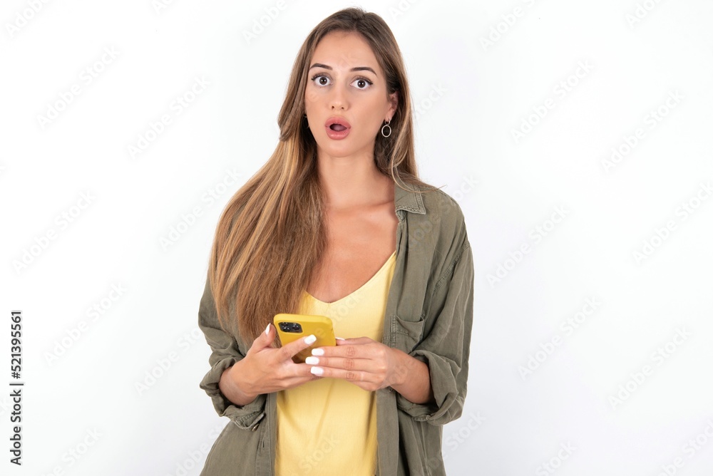 Shocked young beautiful woman wearing green overshirt over white background opens mouth hold phone reading advert unbelievable big shopping prices Shocked young beautiful woman wearing green overshirt over white background opens mouth hold phone reading advert unbelievable big shopping prices