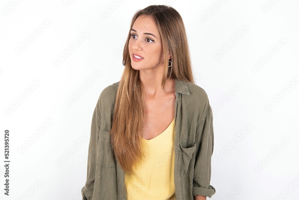 young beautiful woman wearing green overshirt over white background stares aside with wondered expression has speechless expression. Embarrassed model looks in surprise