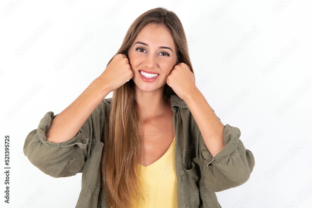Happy young beautiful woman wearing green overshirt over white background keeps fists on cheeks smiles broadly and has positive expression being in good mood Happy young beautiful woman wearing green overshirt over white background keeps fists on cheeks smiles broadly and has positive expression being in good mood