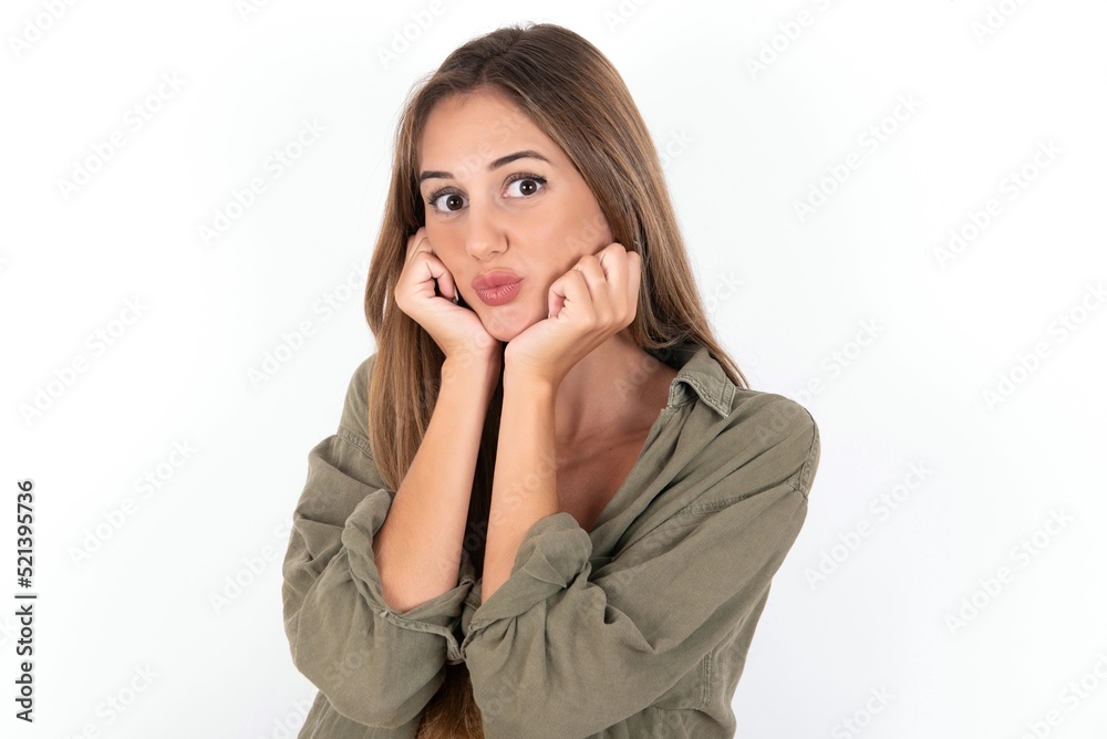 young beautiful woman wearing green overshirt over white background with surprised expression keeps hands under chin keeps lips folded makes funny grimace young beautiful woman wearing green overshirt over white background with surprised expression keeps hands under chin keeps lips folded makes funny grimace