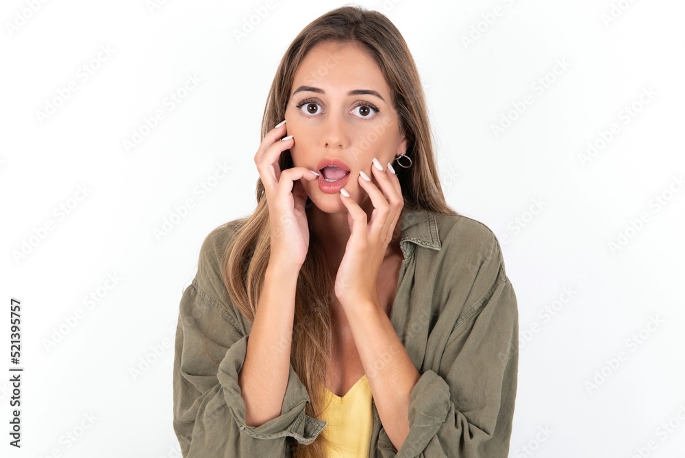 Speechless young beautiful woman wearing green overshirt over white background  keeps hands near opened mouth reacts to shocking news stares wondered at camera