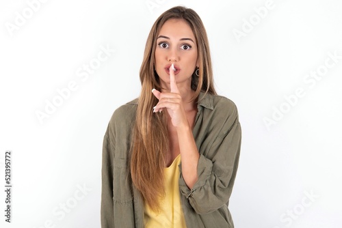 Surprised young beautiful woman wearing green overshirt over white background makes silence gesture  keeps finger over lips and looks mysterious at camera