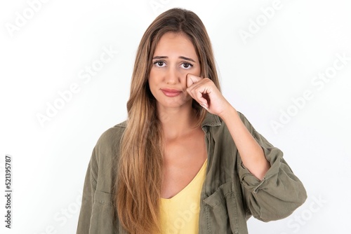 Disappointed dejected young beautiful woman wearing green overshirt over white background wipes tears stands stressed with gloomy expression. Negative emotion