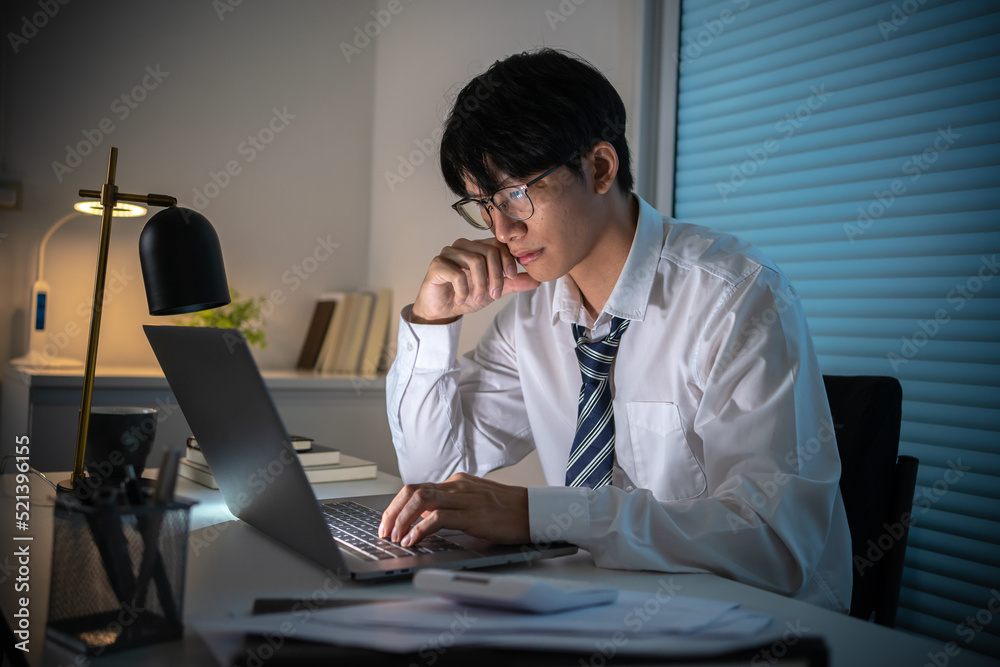 Alone Working Late at Night.Asian business man at workplace in office holding his head on hands after late night work at a desk in a dark office working on a laptop late in the evening concept Alone Working Late at Night.Asian business man at workplace in office holding his head on hands after late night work at a desk in a dark office working on a laptop late in the evening concept