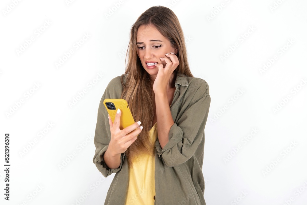 Afraid funny young beautiful woman wearing green overshirt over white background holding telephone and bitting nails Afraid funny young beautiful woman wearing green overshirt over white background holding telephone and bitting nails