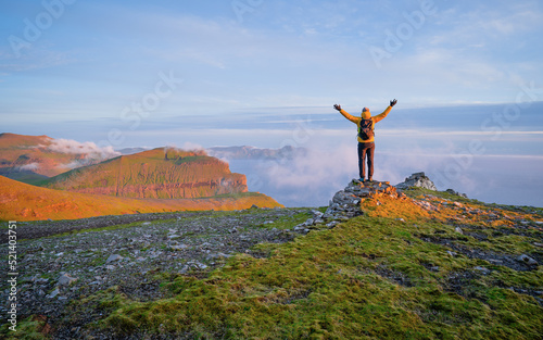 Cheering young man hiker open arms at mountain peak. Beautiful sunset in the mountains. Traveler with raised hands standing on mountain with white fog below and ocean in the background