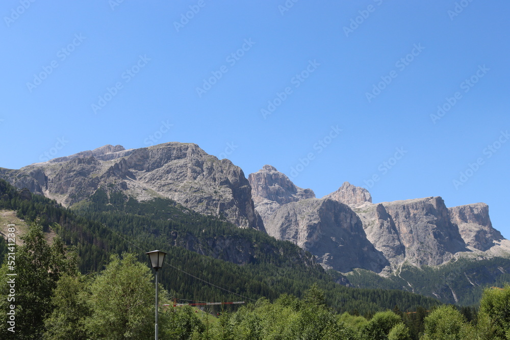 Fototapeta premium Coravara, Italy-July 16, 2022: The italian Dolomites behind the small village of Corvara in summer days with beaitiful blue sky in the background. Green nature in the middle of the rocks. 