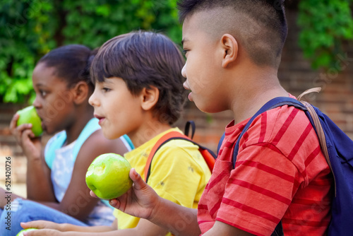 multiethnic kids with backpacks sitting on the street at school entrance eating apples and talking friendly. Healthy eating for children 