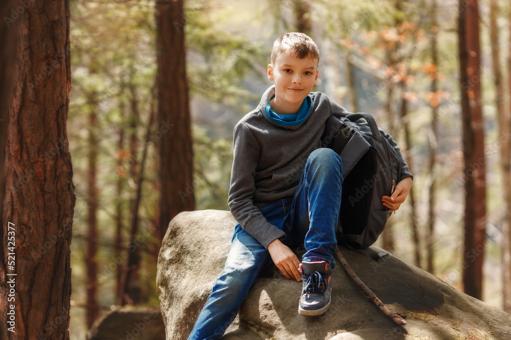 A boy sits on a large stone in nature. Kid is smiling and looking at camera, backpack is near the person. Child has a good mood during autumn sunny day.