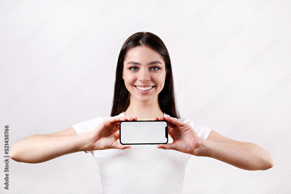 Young beautiful brunette woman with long hair wearing white t-shirt, posing with a phone over isolated background. Portrait of female model showing blank screen cellphone. Close up, copy space Young beautiful brunette woman with long hair wearing white t-shirt, posing with a phone over isolated background. Portrait of female model showing blank screen cellphone. Close up, copy space