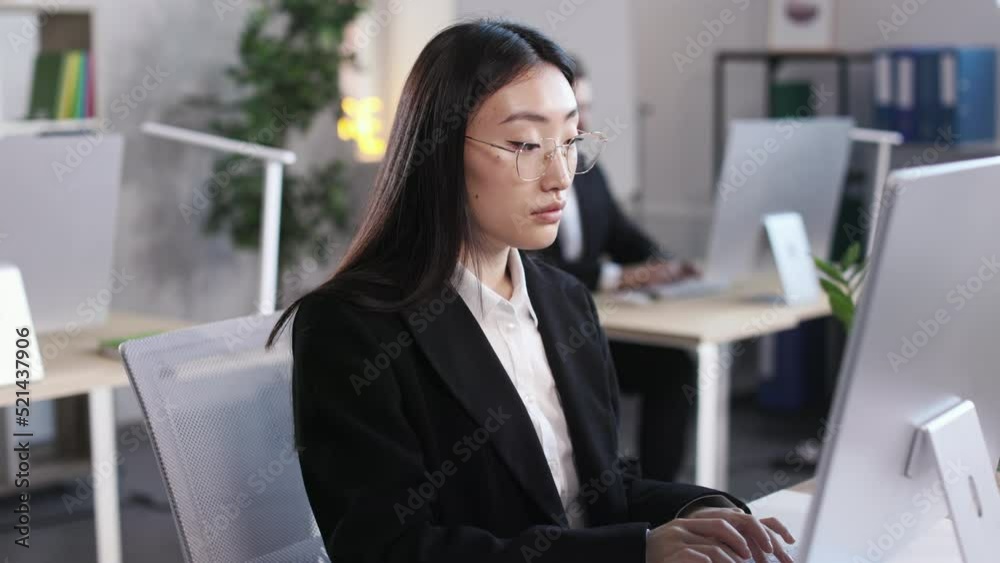 Two multiracial colleagues sitting at desk in office and typing on keyboard of desktop computer. Focus on asian woman in glasses and stylish black suit looking at camera. Two multiracial colleagues sitting at desk in office and typing on keyboard of desktop computer. Focus on asian woman in glasses and stylish black suit looking at camera.