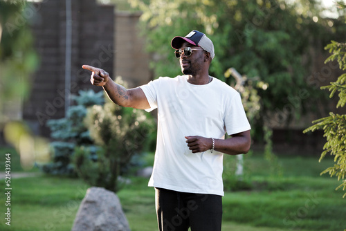 Portrait of African American man in white mockup T-shirt and sunglasses points his finger somewhere outdoor