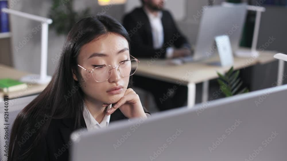Serious asian businesswoman in glasses looking on computer monitor with male colleague on blur background. Young lady in formal wear sitting at desk in office. Serious asian businesswoman in glasses looking on computer monitor with male colleague on blur background. Young lady in formal wear sitting at desk in office.