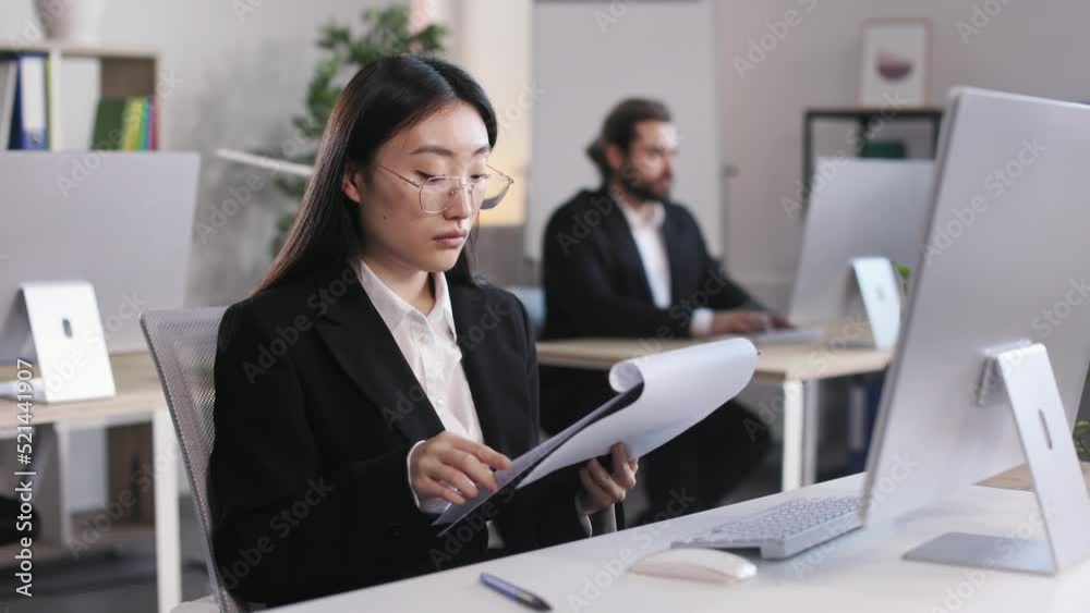 Beautiful asian woman in stylish formal wear sitting at workplace with modern pc and looking through documents on clipboard. Caucasian male colleague working on blur background. Beautiful asian woman in stylish formal wear sitting at workplace with modern pc and looking through documents on clipboard. Caucasian male colleague working on blur background.