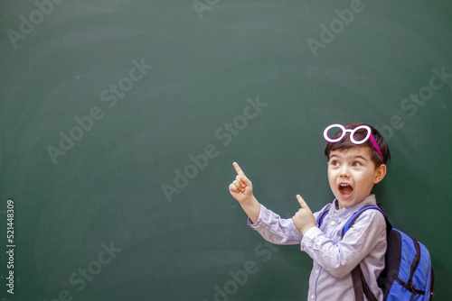 happy kid with party decorative glasses pointing indicates on empty no text green color blackboard .smiling child pointing with hand finger at empty space, raised hands hooray emotion.back to school  photo