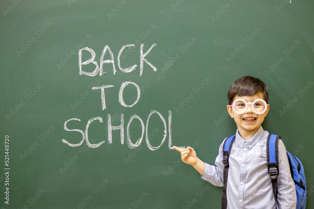 cute little boy preschooler is writing on blackboard with white chalk the words back to school.kid is wearing funny round party glasses with no glass inside.education,learning,classroom interior cute little boy preschooler is writing on blackboard with white chalk the words back to school.kid is wearing funny round party glasses with no glass inside.education,learning,classroom interior