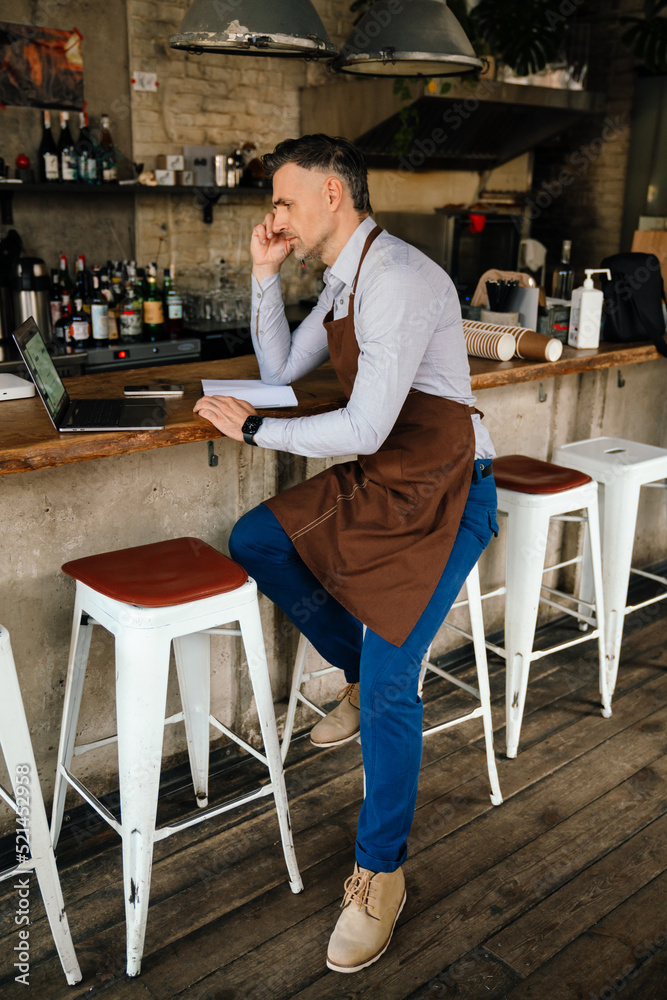 Adult white bearded waiter using laptop while working in cafe indoors Adult white bearded waiter using laptop while working in cafe indoors