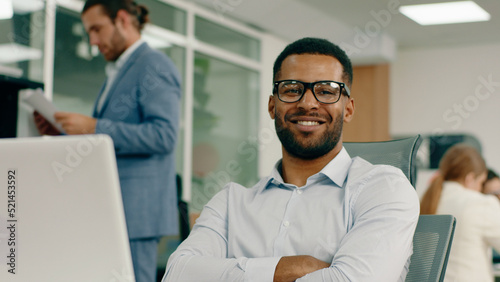 A young man is looking very alluring, he has a beard and glasses, he s in a large office room that is very bright, as he sits back in his chair and crosses his arms smilin